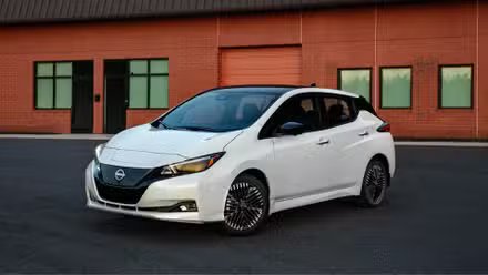 Vue 3/4 avant d'une Nissan Leaf 2025 blanche garée sur le tarmac à l'extérieur d'un bâtiment en brique.