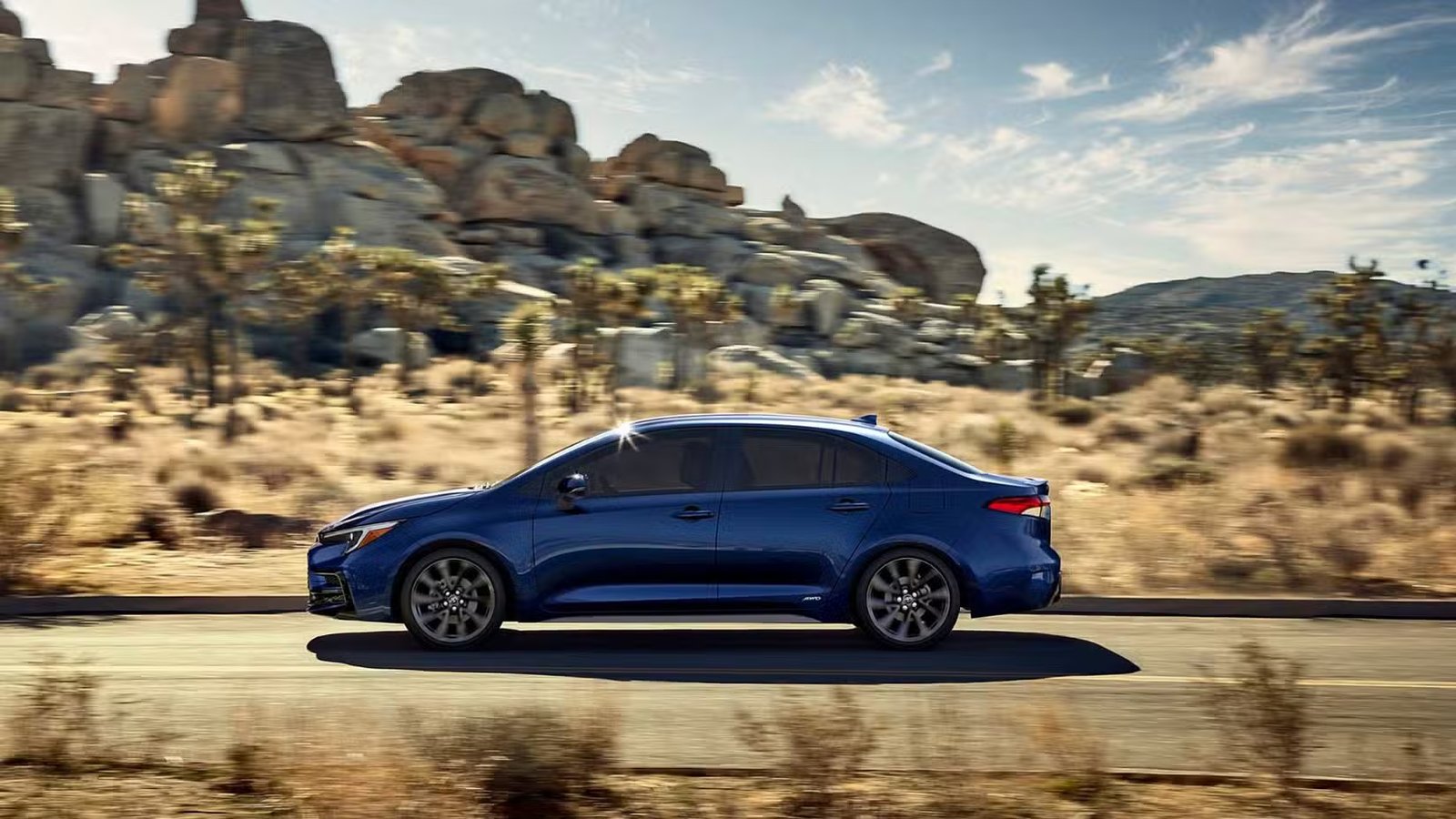 Photo de profil latérale dynamique d'une Toyota Corolla hybride 2025 bleue roulant sur une route de campagne à travers le désert.