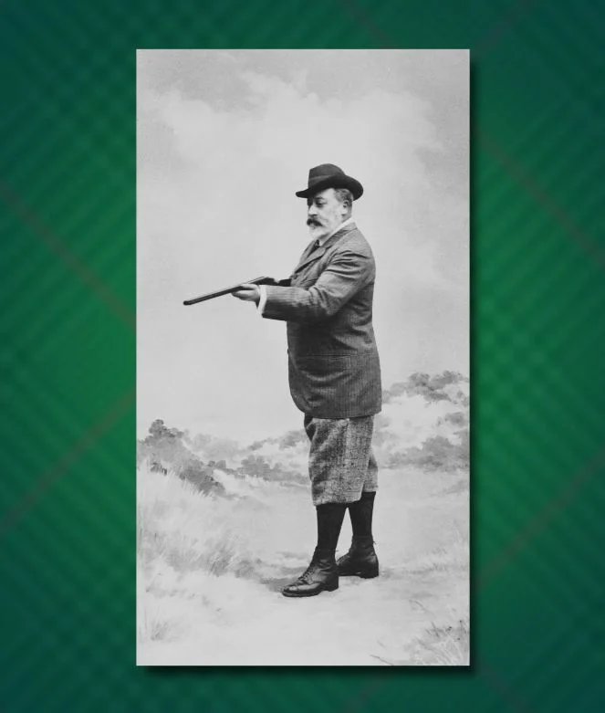 Une photographie vintage en noir et blanc du roi Édouard VII debout dans un champ lors d'une expédition de chasse. Il est représenté de profil, portant un chapeau Homburg, une veste en tweed à motifs et une culotte jusqu'aux genoux avec des chaussettes hautes et des bottes à lacets.