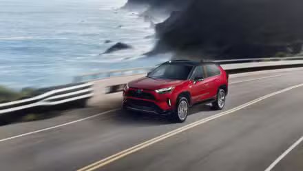 Plan dynamique 3/4 avant d'un Toyota RAV4 Prime XSE 2024 rouge roulant sur une route longeant l'océan.