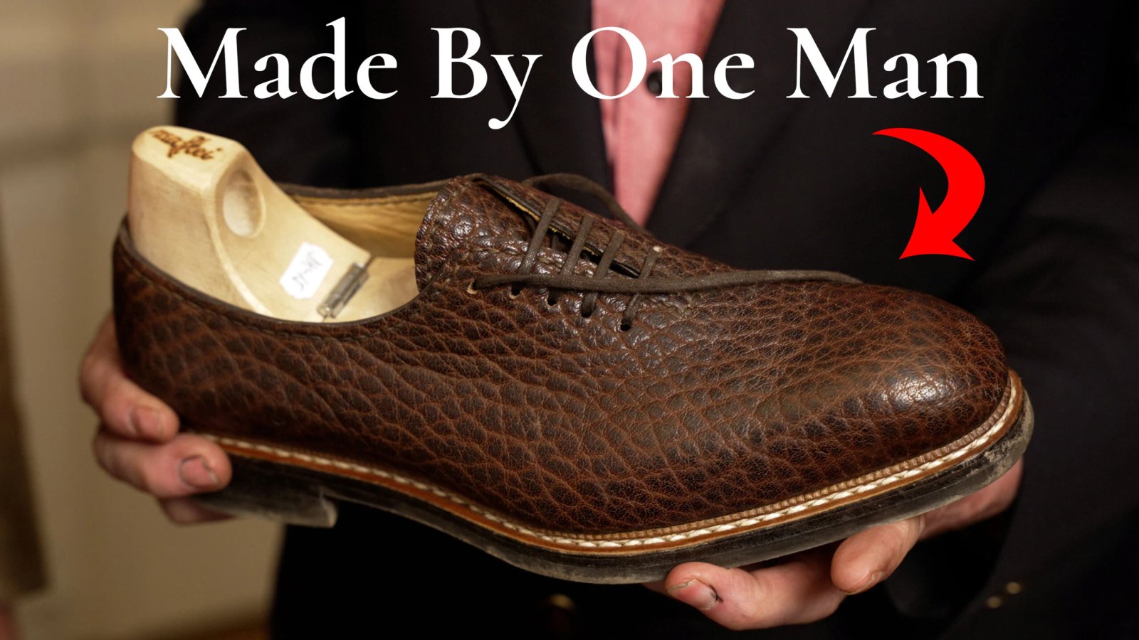 Chaussure habillée en cuir texturé marron faite à la main avec un embauchoir en bois tenu par un artisan ; le texte dit : « Fabriqué par un seul homme. »