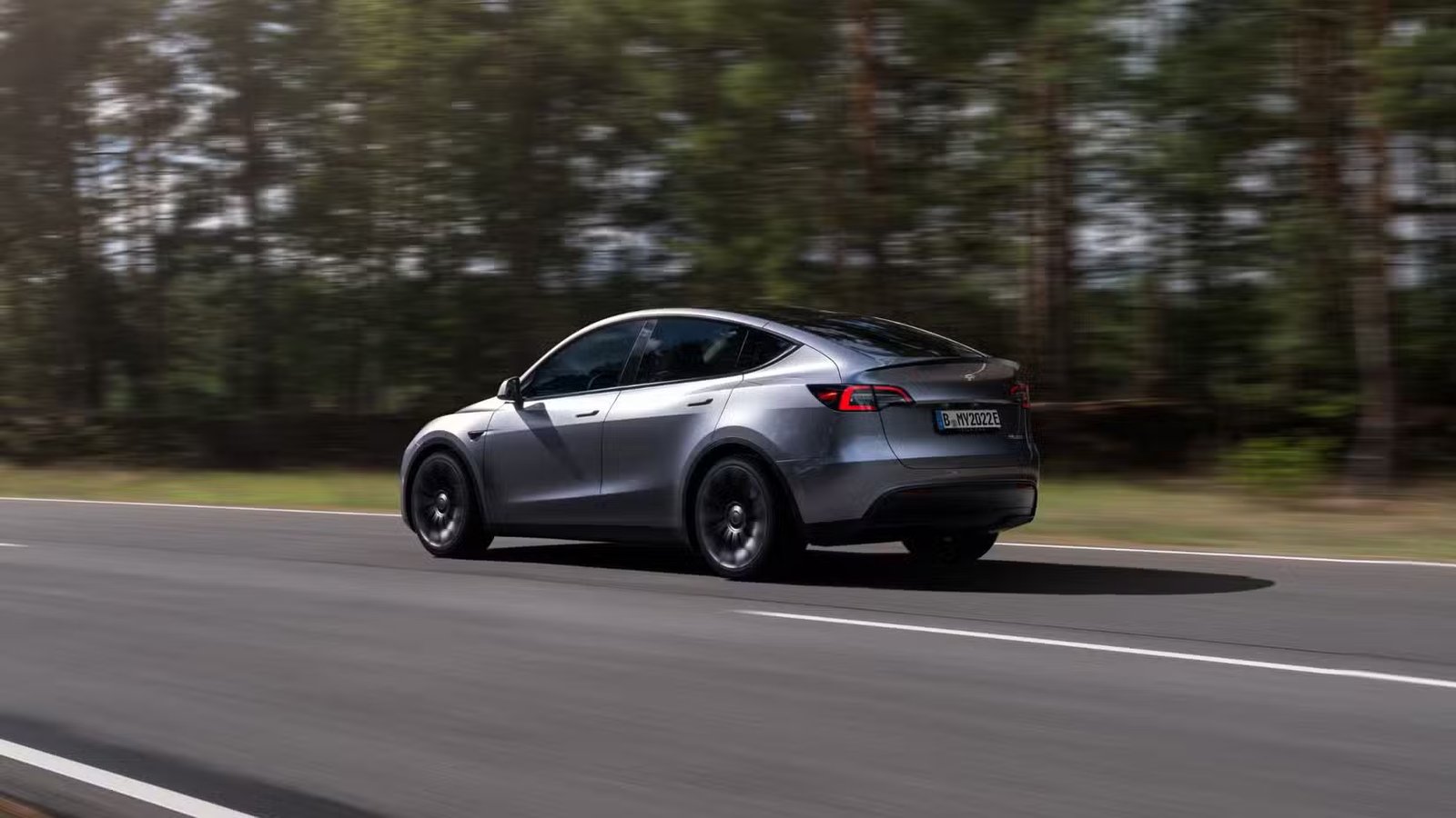 Une Tesla Model Y argentée roulant sur une route de campagne à travers une forêt.