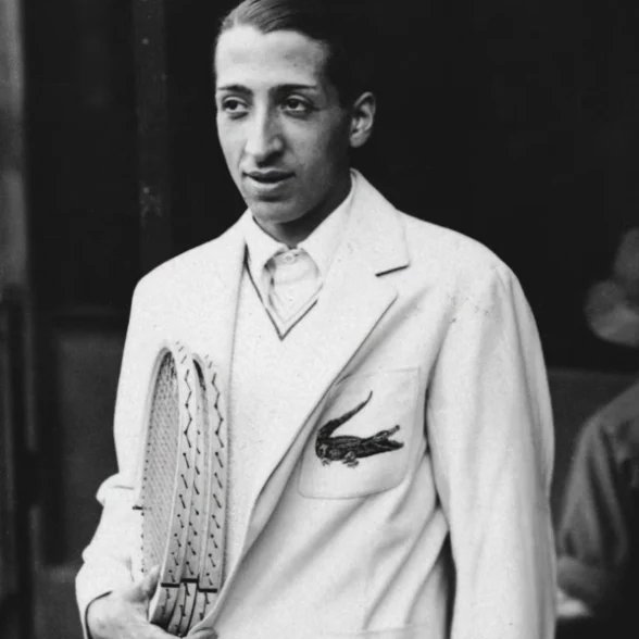 René Lacoste Blazer Crocodile Photo en noir et blanc de René Lacoste dans un blazer de tennis blanc avec un ancien emblème de crocodile sur la poche poitrine, tenant des raquettes de tennis