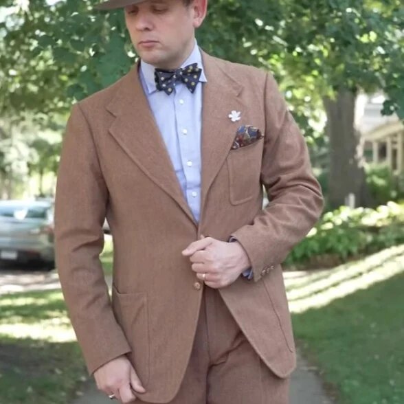 Un homme en extérieur portant un costume en flanelle marron clair avec des poches plaquées, associé à une chemise bleu clair et un nœud papillon à motifs.