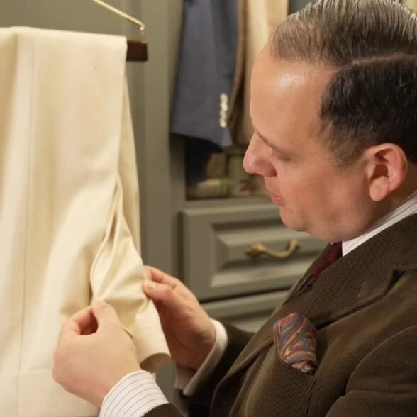 Raphaël inspecte méticuleusement les revers pliés d'un pantalon en laine blanc cassé dans un placard pour vérifier s'il y a des signes d'œufs de mites ou d'activité larvaire.