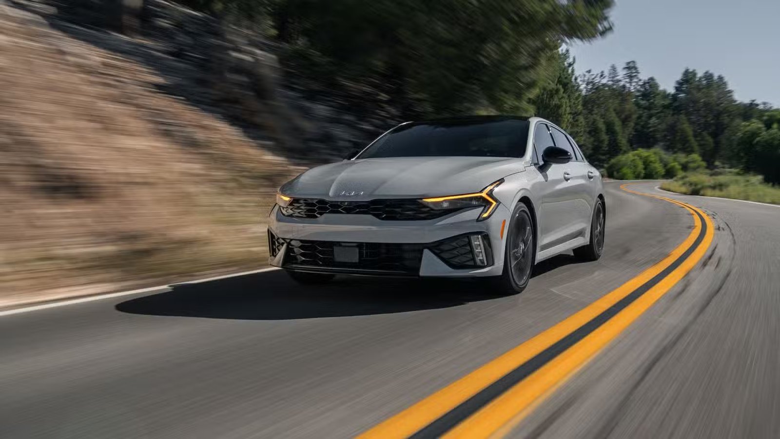 Plan dynamique 3/4 avant d'une Kia K5 2025 grise roulant sur une route de campagne à travers les montagnes.