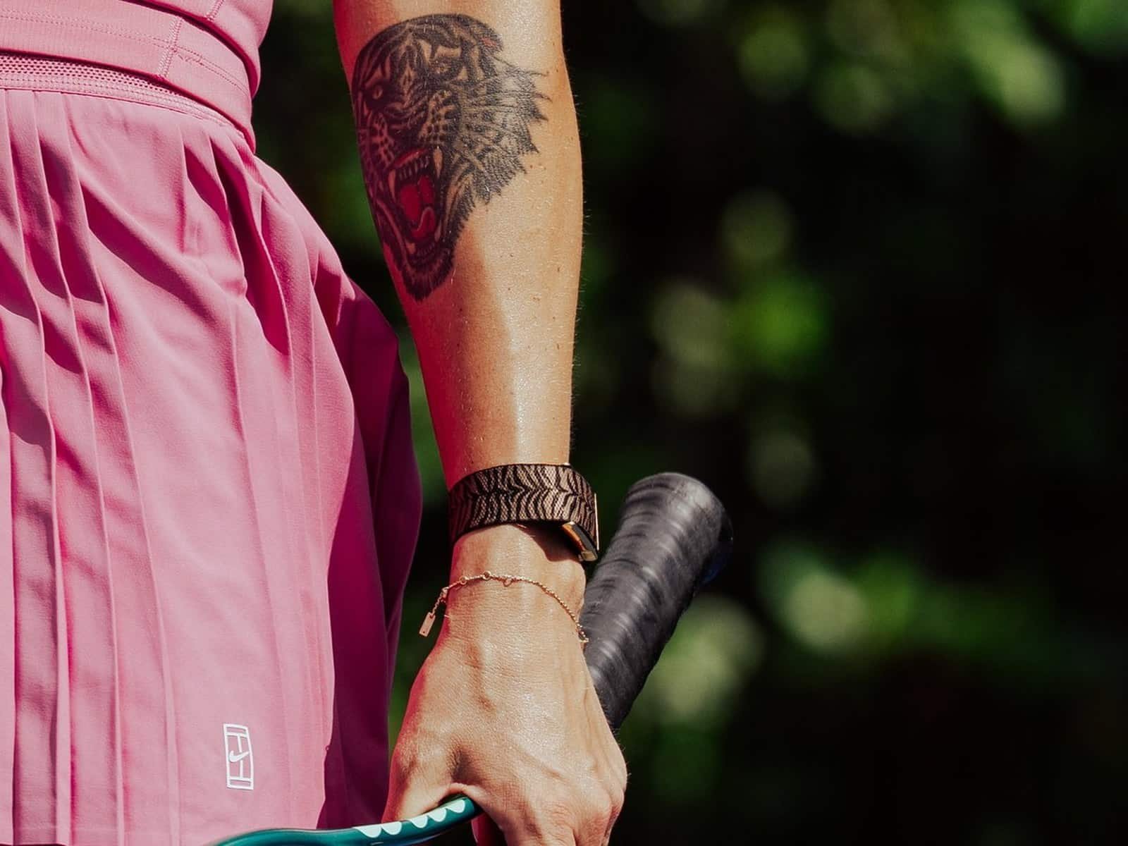 Gros plan de l'avant-bras d'Aryna Sabalenka montrant son tatouage de tête de tigre au-dessus du bracelet WHOOP à rayures tigrées sur son poignet, tenant une raquette de tennis.