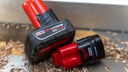 Deux batteries Milwaukee M12 posées sur une scie à table avec de la sciure de bois et une lame en arrière-plan.
