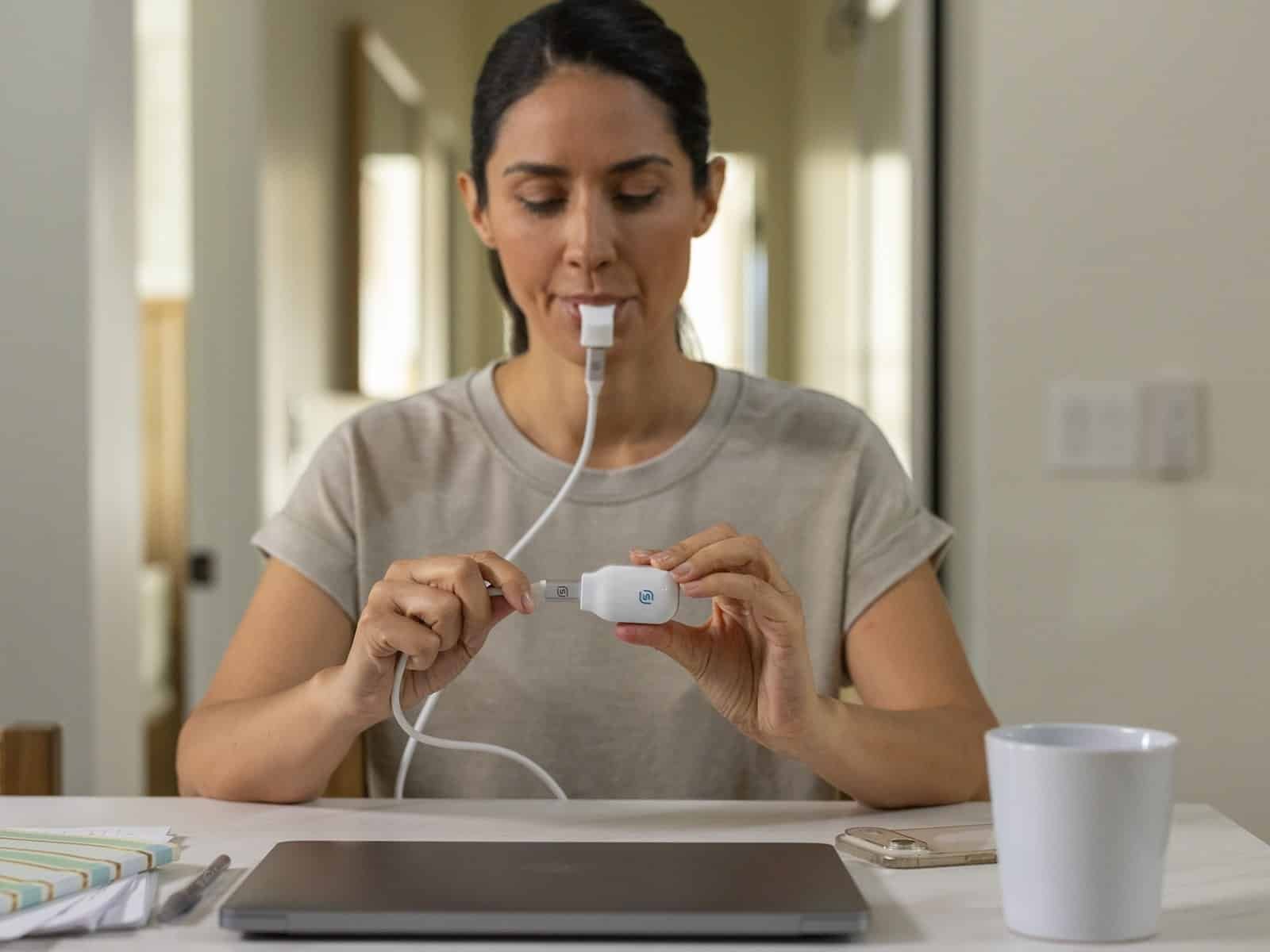 Une femme exécutant une session eXciteOSA de jour sur le banc de sa cuisine, avec l'embout buccal connecté au contrôleur portable.