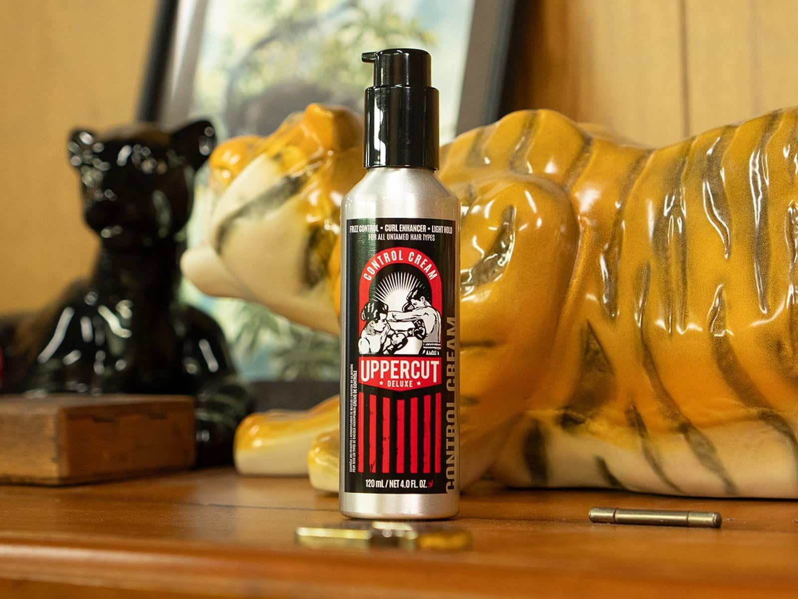 Uppercut Deluxe Control Cream debout sur une commode en bois devant un ornement de tigre en céramique et une figurine de panthère noire.