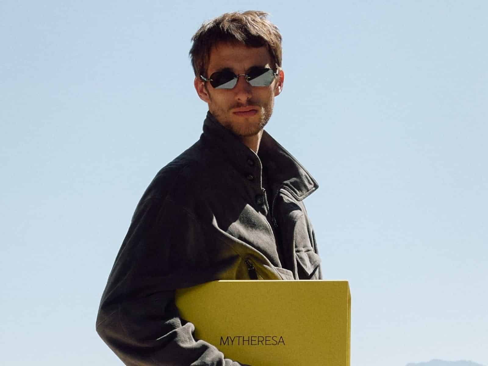 Un mannequin portant des lunettes de soleil et une veste militaire en daim foncé tenant une boîte Mytheresa jaune, avec des montagnes derrière lui.