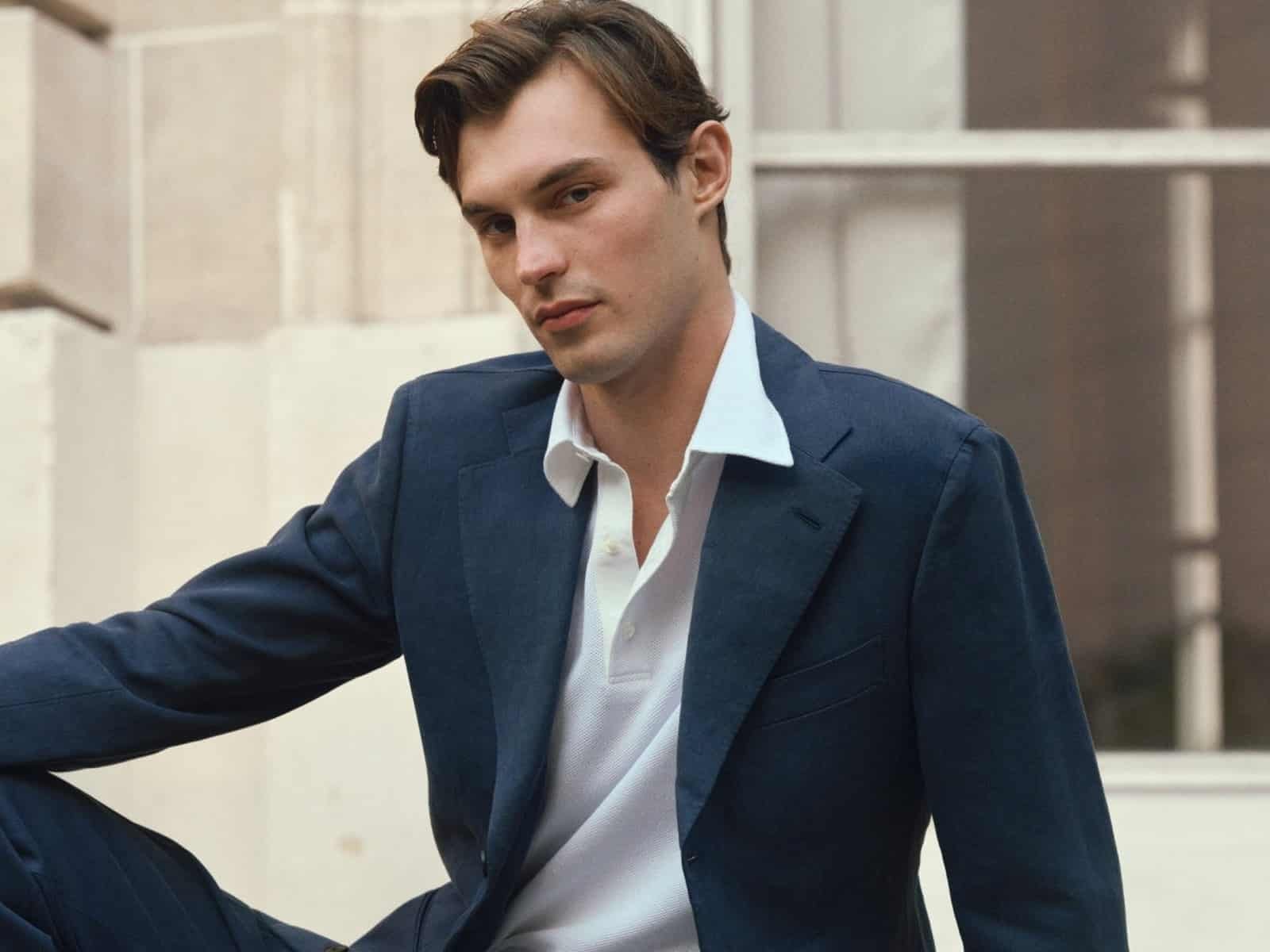 Un mannequin vêtu d'un costume bleu marine et d'un polo piqué blanc, assis sur des marches en pierre à l'extérieur d'un bâtiment en pierre.