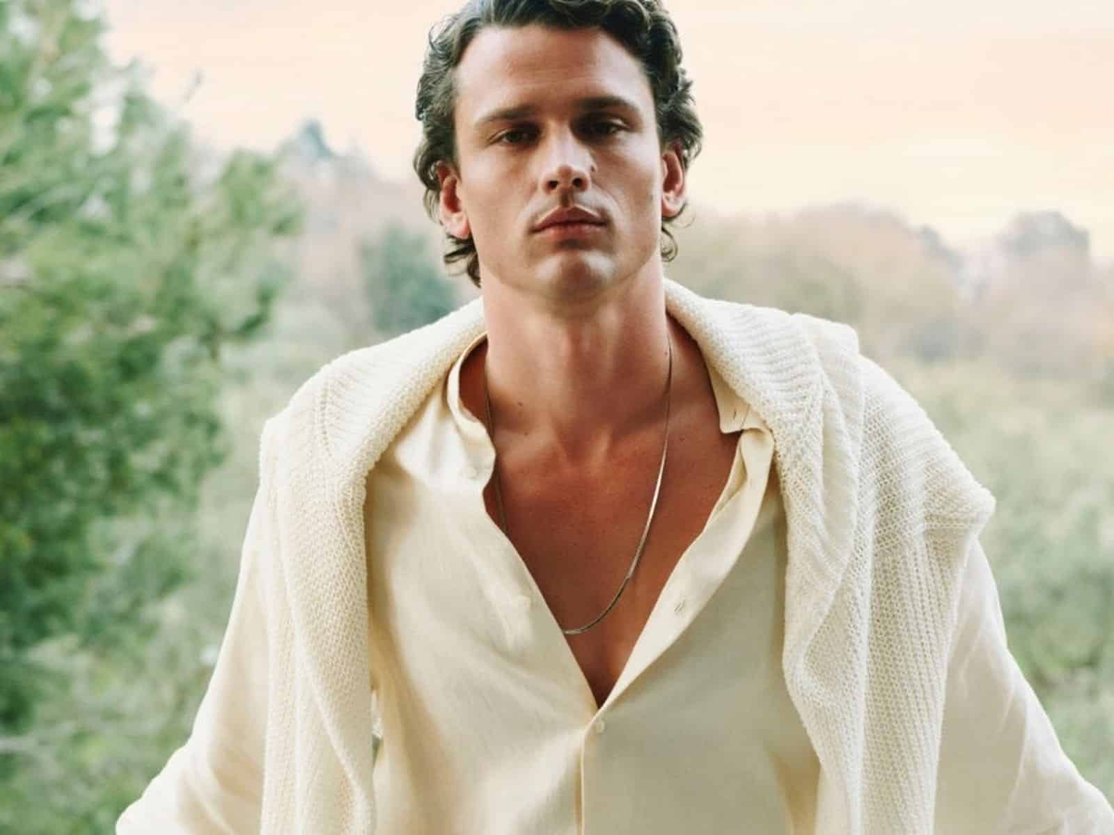 Un mannequin vêtu d'une chemise crème à col ouvert avec un tricot crème drapé sur les épaules, debout devant des oliviers au crépuscule.