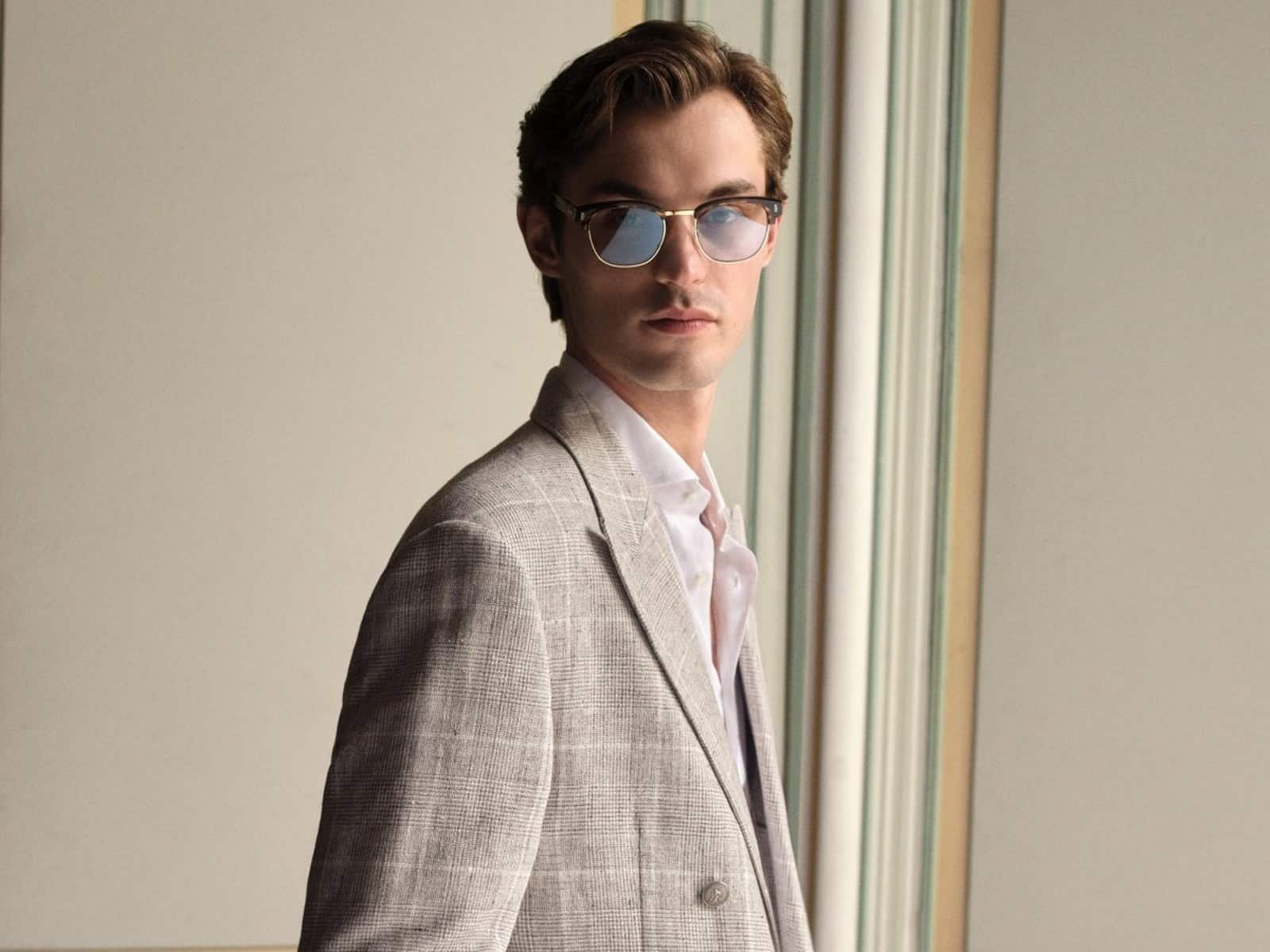 Un mannequin portant un blazer gris à carreaux Prince de Galles et une chemise blanche à col ouvert, debout à côté des portes à panneaux.