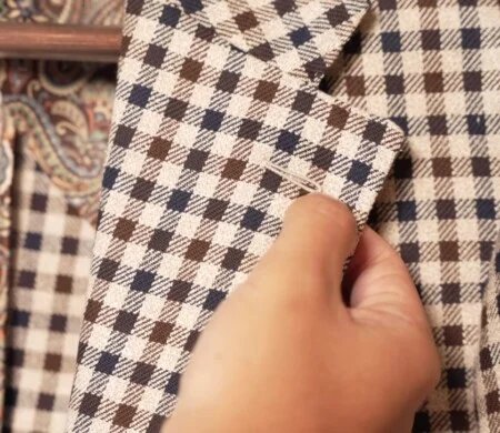 Vidéo Highlight 2 - Veste à carreaux avec boutonnière et revers Une main indiquant la boutonnière du revers d'une veste à carreaux beige, bleu marine et marron, vue de près