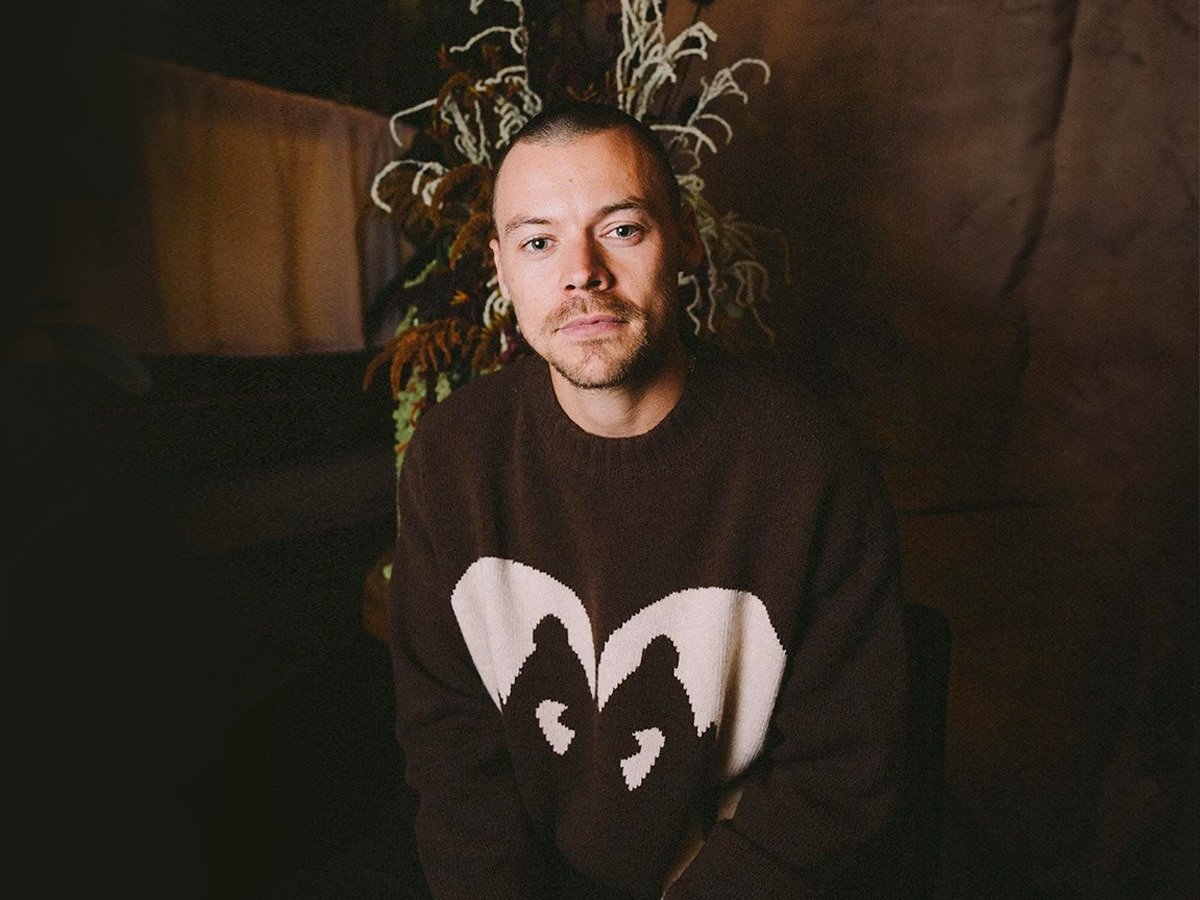 Homme à la coupe buzz portant un pull marron à motif blanc, assis à l'intérieur avec des plantes en arrière-plan.