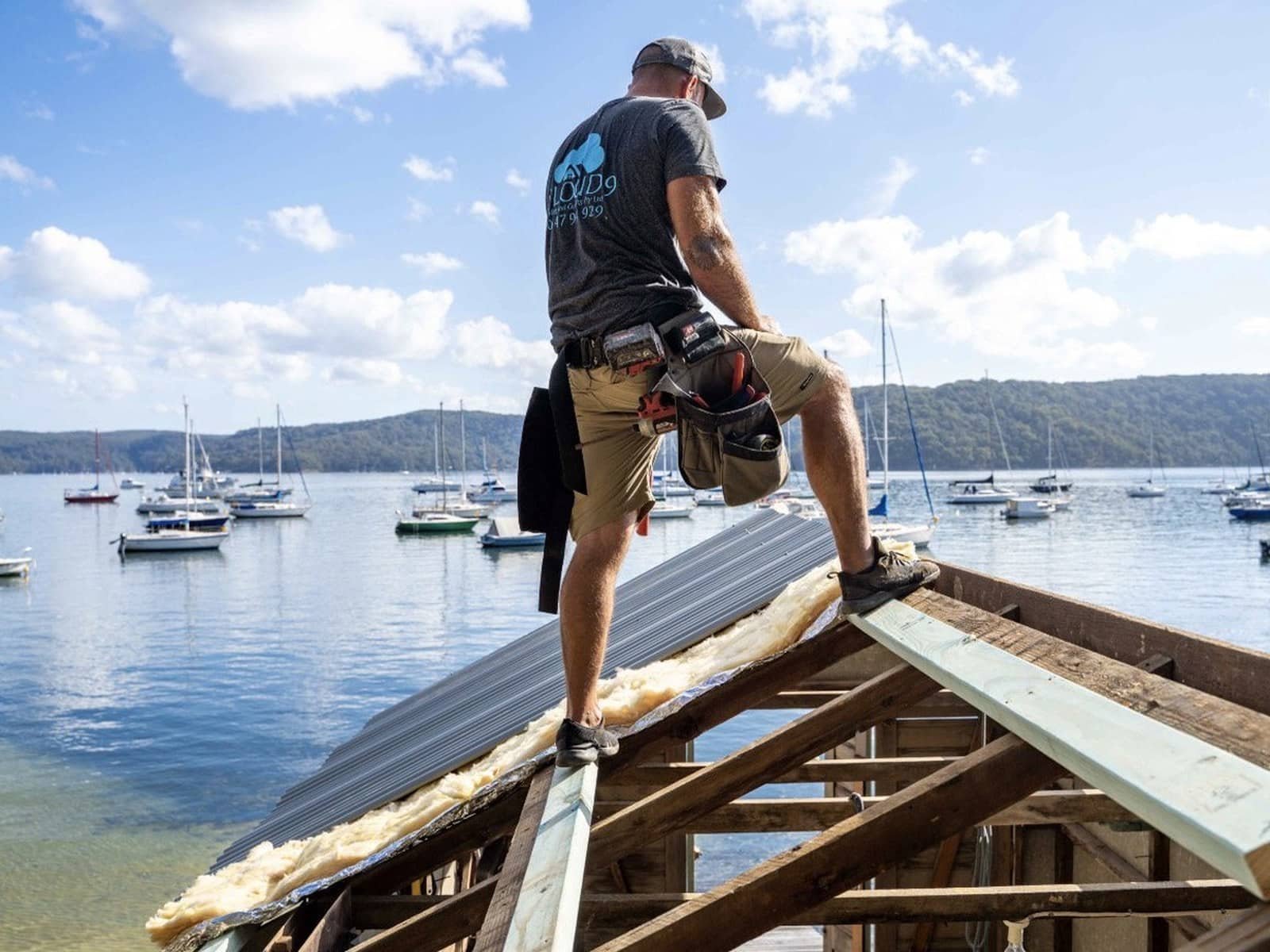 Tommy Alsop en short de travail et une ceinture à outils debout sur une charpente de toit exposée au-dessus d'une baie pleine de bateaux amarrés