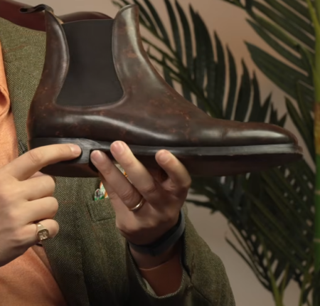 Vidéo d'examen des bottines Chelsea, point culminant 2 Vue rapprochée d'une botte Chelsea en cuir marron foncé patiné tenue à deux mains, montrant le panneau latéral élastique, le bout en amande et la taille biseautée