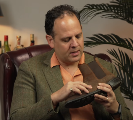 Examen des bottes Chelsea Raphael Raphael, vêtu d'une veste en tweed à chevrons verts et d'un polo orange, examine la semelle et le talon d'une botte Chelsea en daim marron alors qu'il est assis dans un fauteuil en cuir.