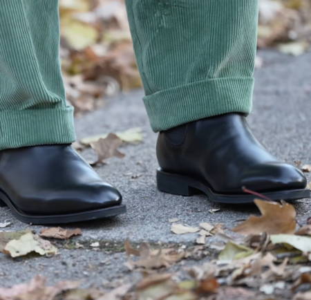 Vidéo d'examen des bottines Chelsea, point culminant 3 Bottines Chelsea en cuir noir portées avec un pantalon en velours côtelé vert sauge à revers, photographiées en extérieur sur un trottoir parsemé de feuilles d'automne