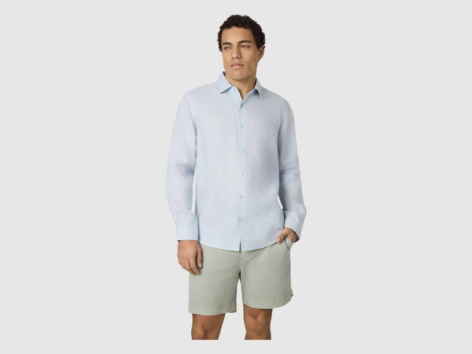 Un homme portant la chemise en lin Connor Hamilton en bleu clair avec un short sauge, une main dans sa poche