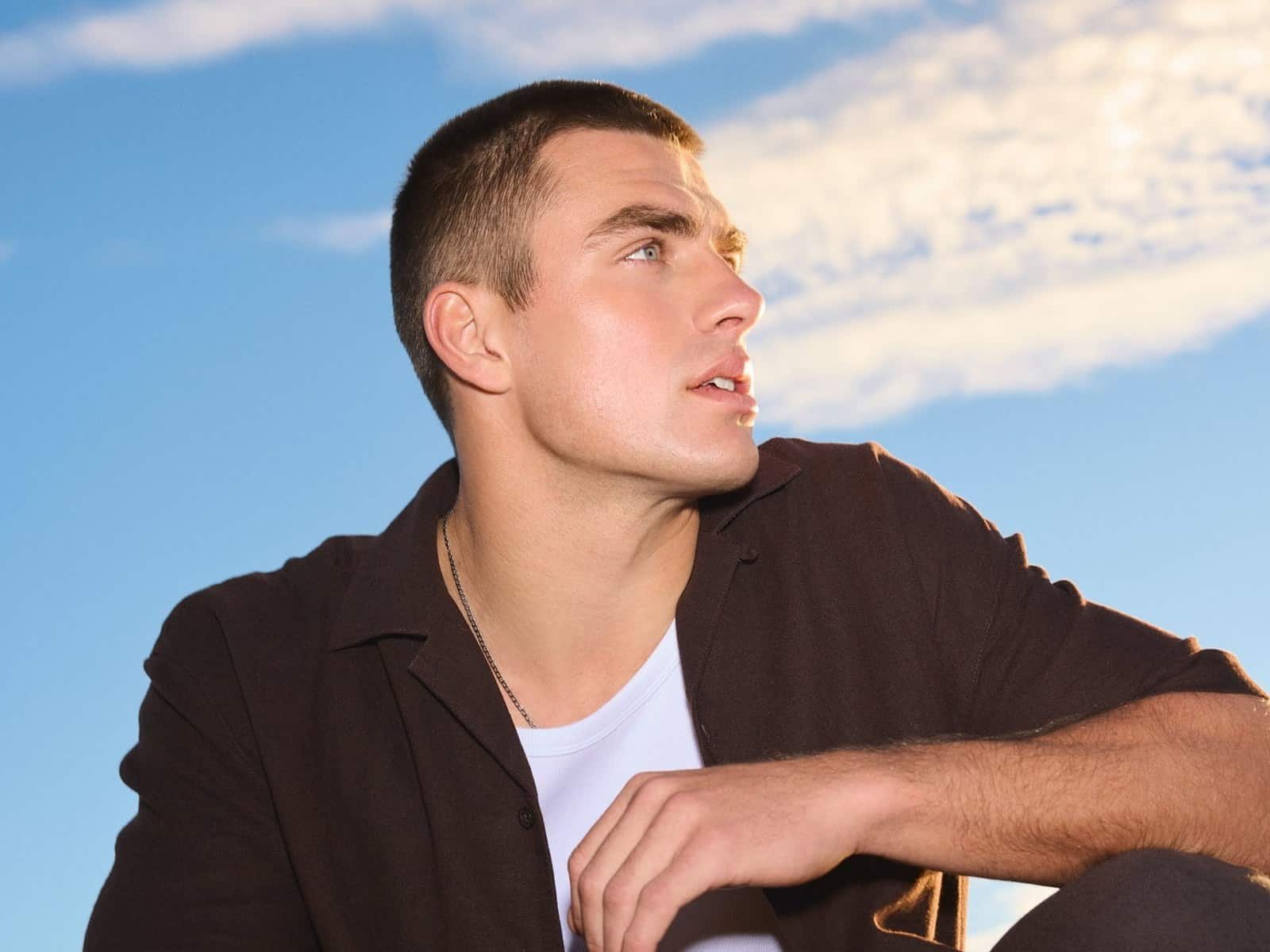 Un homme vêtu d'une chemise sombre à manches courtes sur un t-shirt blanc, accroupi contre un ciel bleu vif
